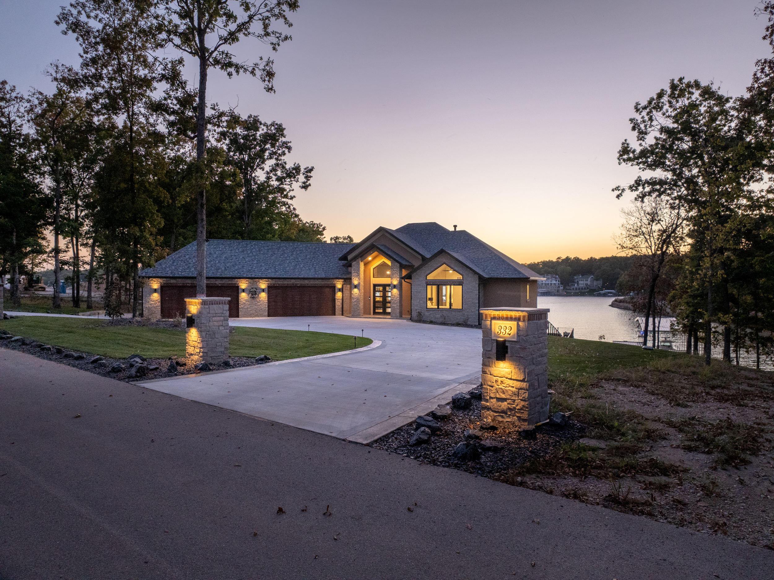 Custom single-level lakefront stone home at dusk with lit stone entry pillars